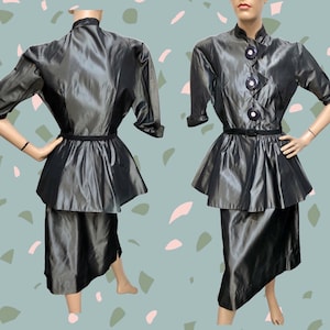 Op de afbeelding: Een vintage tweedelig pak in een donkergrijze, bijna zwarte kleur. De jas heeft een hoge kraag, drie grote knopen en een peplum taille. De rok is knielang met een asymmetrische zoom. Het pak wordt op een mannequin getoond.