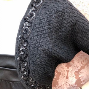 May include: Black knitted shrug with a sequined trim around the neckline. The sequins are black and form a decorative pattern.