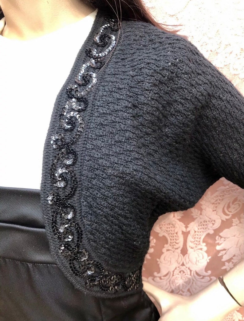 May include: Black knitted shrug with a sequined trim around the neckline. The sequins are black and form a decorative pattern.