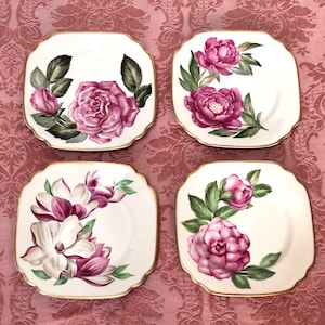 May include: Set of four square ceramic plates with gold trim. Each plate features a different floral design with pink and white flowers on a white background.