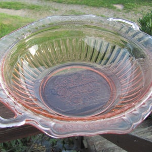 Könnte beinhalten: Eine Vintage-Servierschüssel aus rosa Glas mit gewelltem Rand und Griff. Die Schüssel hat ein geripptes Innendesign und ein Blumenmuster auf der Basis. Die Schüssel besteht aus klarem Glas mit rosa Farbton.