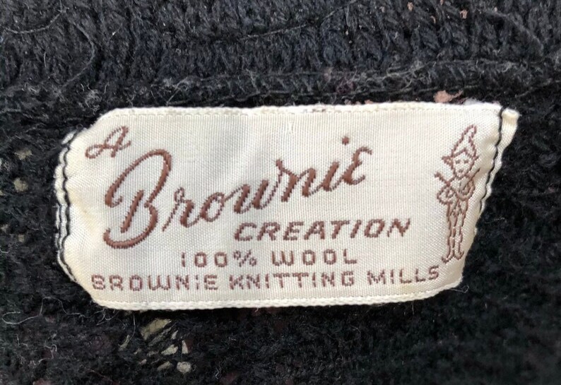 May include: A white label with brown text that reads "A Brownie Creation 100% Wool Brownie Knitting Mills". A cartoon illustration of a person holding a musical instrument is on the right side of the label.