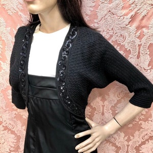 May include: Black knitted shrug with a beaded trim. The shrug has a short sleeve and a scoop neck.