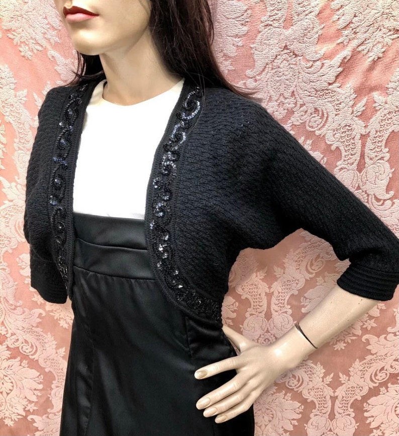 May include: Black knitted shrug with a beaded trim. The shrug has a short sleeve and a scoop neck.