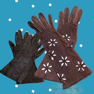 May include: A pair of dark brown gloves featuring white floral cut-out patterns on the cuffs and back of the hands, paired with a second pair of plain dark brown gloves. The gloves have visible white stitching along the fingers and seams.