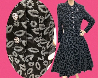 May include: Black vintage dress with a floral pattern and long sleeves. The dress features a button-up front and a flared skirt. The close-up shows the floral embroidery and the decorative buttons. A brooch is pinned on the collar.