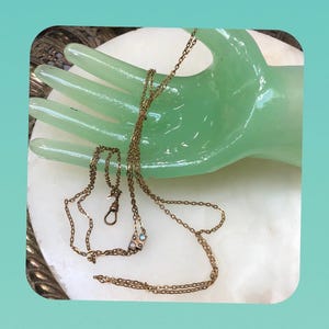 May include: A gold-toned metal chain necklace featuring a delicate link design. The necklace includes a small rectangular pendant set with four tiny, iridescent cabochon stones and a classic swivel clasp mechanism at the end of the chain.