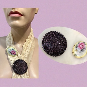 May include: Two vintage brooches, one with a purple rhinestone design and the other with a floral design on a white background.