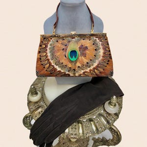 May include: A rectangular handbag featuring a prominent peacock feather eye at the center, surrounded by layered brown and tan feathers. The bag has a gold-tone metal frame, a top clasp closure, and a thin brown handle. A pair of dark brown gloves is included.