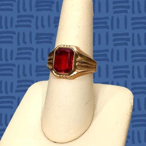 May include: A gold-toned ring featuring a large, rectangular, faceted red gemstone set in the center. The band of the ring displays a series of parallel, grooved lines extending from the central setting along the sides of the metal shank.