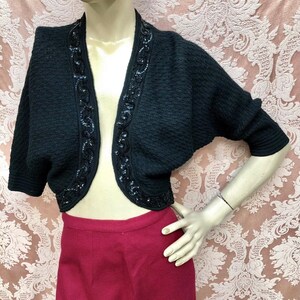 May include: A black cropped cardigan with a beaded trim. The cardigan has a V-neck and short sleeves.