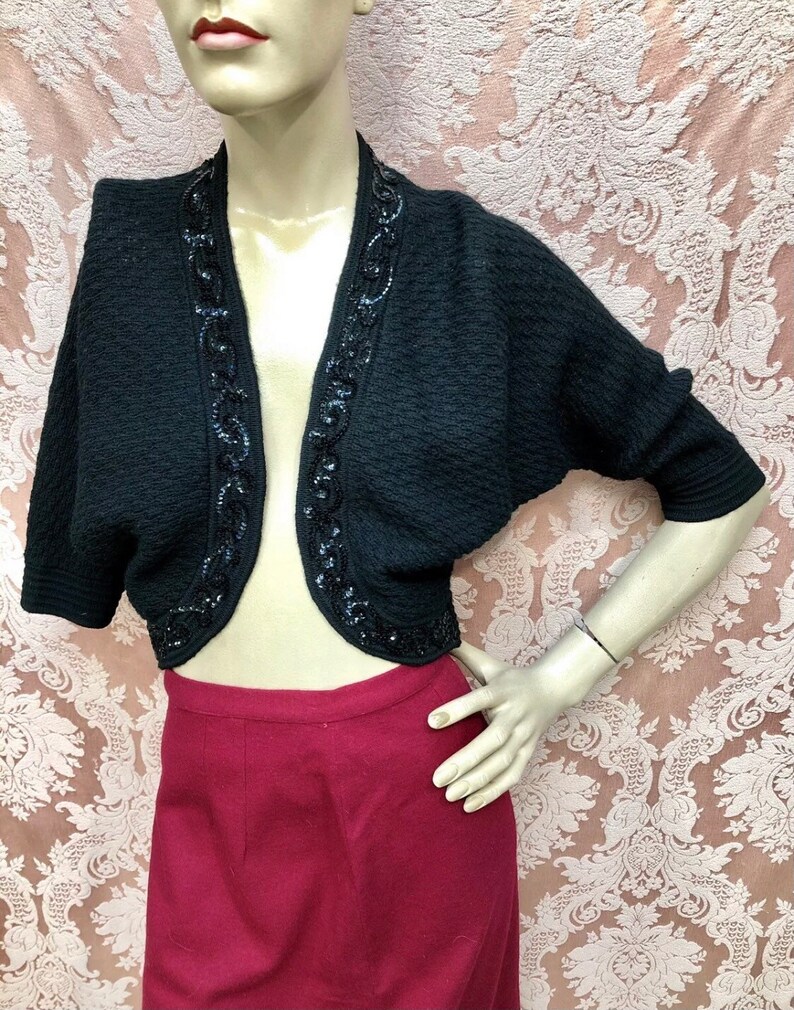 May include: A black cropped cardigan with a beaded trim. The cardigan has a V-neck and short sleeves.
