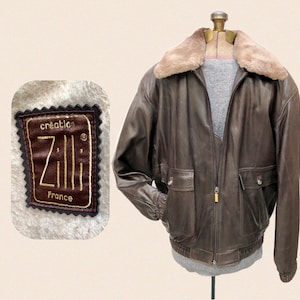 May include: Brown leather jacket with a tan faux fur collar. The jacket has two front pockets and a zipper closure. The label reads "création Zilli France". The jacket is displayed on a mannequin.