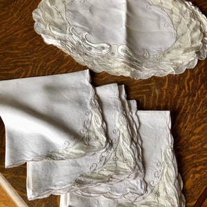 May include: Set of four white linen napkins with delicate embroidered floral designs in a light gray color. The napkins have scalloped edges and are folded neatly.