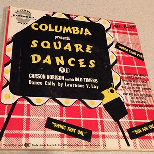 May include: A red and white checkered background with a black sign that reads "Columbia presents Square Dances" with the text "Carson Robison and his Old Timers Dance Calls by Lawrence V. Loy" below. The sign has a yellow border and white light bulbs around the edge. The sign is on a stand with a yellow base and a black pole. The text "Swing That Gal" is in the lower left corner and "Dive for the Oyster" is in the lower right corner.