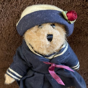 Peut inclure: Un ours en peluche portant une robe de marin bleue avec des bordures blanches, un chapeau de marin blanc et bleu avec une rose rouge et un ruban bordeaux.