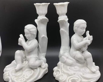 May include: A pair of white porcelain candle holders featuring cherub figures. Each holder has a sculpted base and a trumpet-shaped candle cup.