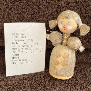 May include: A handmade doll with a straw-like body and a wooden head. The doll has blue eyes, pink cheeks, and a red mouth. The doll is wearing a straw skirt with a geometric pattern. The doll is labeled "ALENKA" and has a price tag attached.