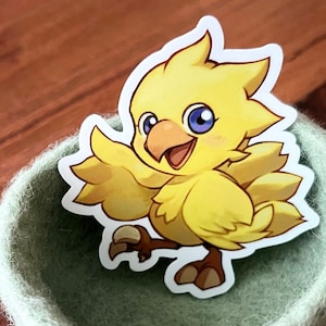 May include: A yellow and white cartoon bird sticker with a blue eye, a yellow beak, and a happy expression. The sticker is placed in a green felt bowl.