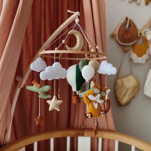 Puede incluir: Un móvil de bebé de madera con un esquema de color marrón claro, verde y blanco. El móvil presenta un globo aerostático, un avión, nubes, estrellas y un gato con gafas de aviador.