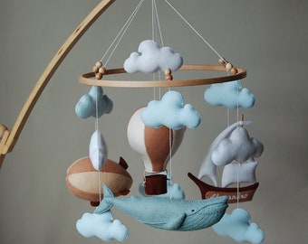 May include: A baby mobile with a wooden frame, displaying felt shapes. The mobile includes a hot air balloon, a whale, a ship, and clouds in blue, brown, and white. The mobile is suspended above a wooden cot.