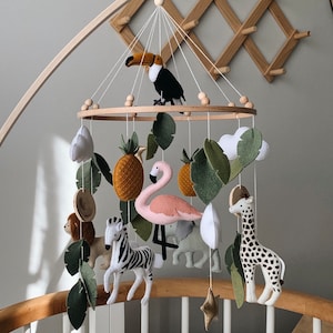 May include: A baby mobile featuring felt animal figures, including a toucan, giraffe, zebra, lion, flamingo, and elephant, along with felt leaves, pineapples, and clouds. The mobile is suspended from a wooden frame.