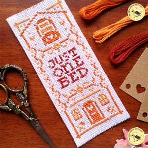 May include: Cross stitch pattern for a bookmark with a house and the text "Just One Bed". The pattern is worked in shades of orange and red on white Aida cloth.