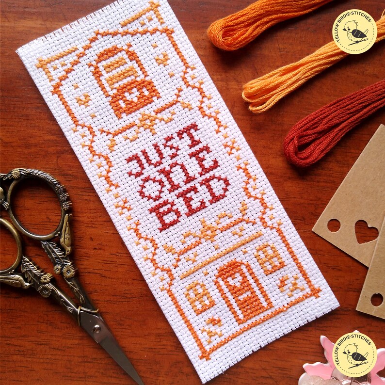 May include: Cross stitch pattern for a bookmark with a house and the text "Just One Bed". The pattern is worked in shades of orange and red on white Aida cloth.