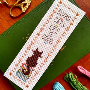 Puede incluir: Un marcapáginas de punto de cruz blanco con un gato negro sentado en una pila de libros. El marcapáginas tiene el texto "Books Cats Life is Good" en hilo marrón y naranja.