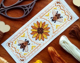 May include: A cross-stitch pattern featuring a sunflower and two bees. The pattern is on white Aida cloth and is surrounded by a brown border. The pattern is surrounded by embroidery floss in various shades of yellow, brown, and white.