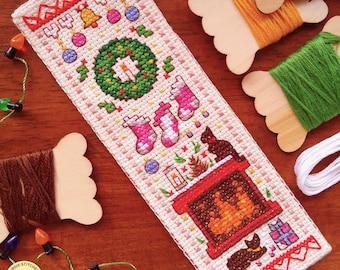 Puede incluir: Un marcapáginas navideño hecho a mano con punto de cruz. El diseño presenta una corona, calcetines, una chimenea con gatos y adornos. El marcapáginas es de color crema con detalles en rojo, verde, rosa y marrón. El texto "Yellow Birdie Stitches" es visible.