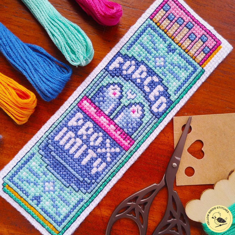 May include: A rectangular cross-stitch bookmark featuring a pixelated design of a blue bottle with the text "F&Uuml;PLED PROX IMIT!!" in white letters. The embroidery uses shades of blue, pink, and teal thread on white fabric, accompanied by embroidery floss.