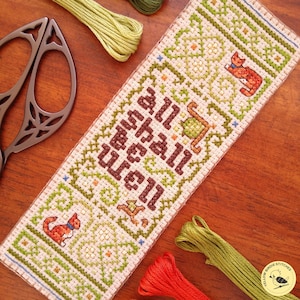 May include: Cross-stitch bookmark with the text "All Shall Be Well" in brown. The bookmark features a green and cream geometric pattern with orange cat illustrations. Embroidery floss and scissors are visible on a wooden surface.