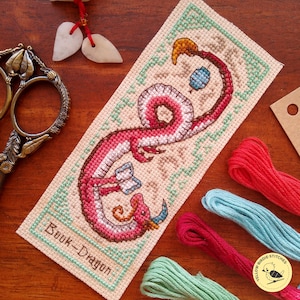 May include: Cross-stitch bookmark featuring a pink and white dragon with a book, the text "Book-Dragon", and a light green border. The bookmark is surrounded by embroidery floss in red, blue, and green.