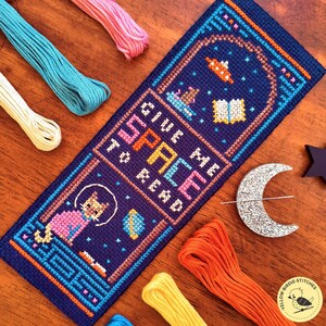 May include: A handmade cross-stitch bookmark with a space theme. The navy blue bookmark features a cat in a spacesuit, stars, and the text "GIVE ME SPACE TO READ" in colorful letters. Embroidery floss in various colors surrounds the bookmark.