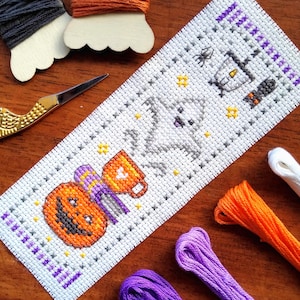 May include: A handmade Halloween cross-stitch bookmark. The white fabric features a smiling jack-o'-lantern, a ghost, a spider, and a candle. Purple and orange embroidery threads and gold scissors are also visible.
