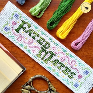 May include: A white cross-stitch bookmark embroidered with the words "Fated Mates" in green. The bookmark is decorated with pink, green, and blue floral and ribbon patterns. Various coloured embroidery threads are arranged around it.