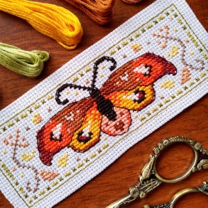 May include: A cross-stitch pattern of a butterfly with orange, yellow, and brown wings. The butterfly is surrounded by a green border with small floral designs.