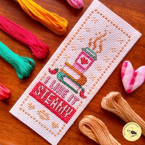 Pode incluir: Um marcador de livros de ponto cruz feito à mão com um desenho de chávena de café e o texto "I LIKE IT STEAMY". O marcador de livros é branco com um desenho de chávena de café rosa, verde e castanho. Fio de bordar colorido em tons de rosa, amarelo e verde rodeia o marcador.