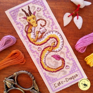 May include: Cross-stitch bookmark featuring a yellow and orange dragon holding a cake, with the text "Cake-Dragon". The bookmark has a lavender border and is surrounded by embroidery floss and scissors.