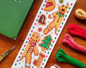 Puede incluir: Un marcapáginas hecho a mano con punto de cruz con hombres de jengibre y galletas. El marcapáginas blanco tiene diseños de punto de cruz marrón, rojo, verde y amarillo. Un libro verde y hilo de bordar en el fondo.
