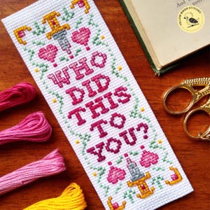 K&ouml;nnte beinhalten: Wei&szlig;er Kreuzstich-Lesezeichen mit gr&uuml;nem und gelbem floralem Rand. Das Lesezeichen zeigt ein rosa Herz, einen Dolch und den Text "I WHO DID THIS TO YOU?"