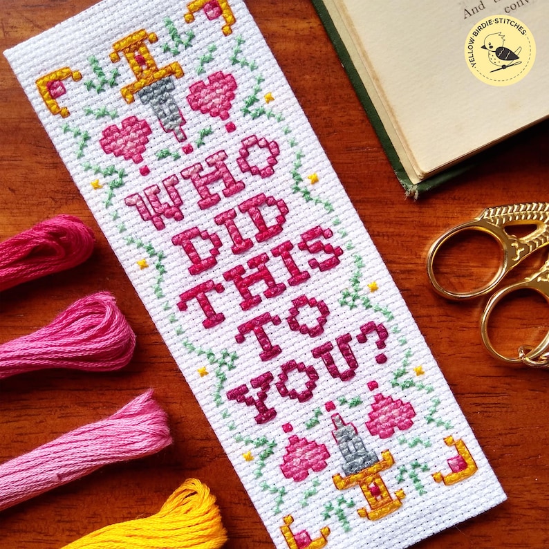 K&ouml;nnte beinhalten: Wei&szlig;er Kreuzstich-Lesezeichen mit gr&uuml;nem und gelbem floralem Rand. Das Lesezeichen zeigt ein rosa Herz, einen Dolch und den Text "I WHO DID THIS TO YOU?"