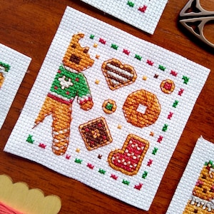 May include: Cross-stitch design showing a gingerbread man and assorted biscuits. The gingerbread man wears a green jumper. Biscuits include a heart, a square, a doughnut, a circle, and a stocking. The design is on white fabric with red, green, and yellow accents.