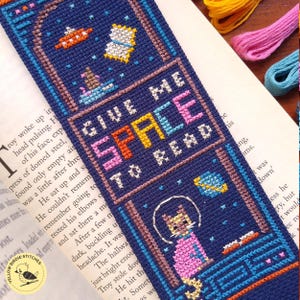 May include: A navy blue cross-stitch bookmark with a space theme. It features a cat in a spacesuit, planets, and the text "GIVE ME SPACE TO READ" in colorful letters. The bookmark is a handmade item.