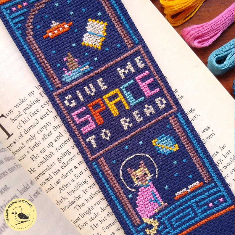 May include: A navy blue cross-stitch bookmark with a space theme. It features a cat in a spacesuit, planets, and the text "GIVE ME SPACE TO READ" in colorful letters. The bookmark is a handmade item.