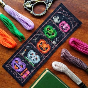 Puede incluir: Un marcapáginas de tela negra con un diseño de punto de cruz que presenta una fila de calabazas coloridas. Las calabazas son naranja, verde, morado y rosa. El marcapáginas está rodeado de hilo de bordar de colores.