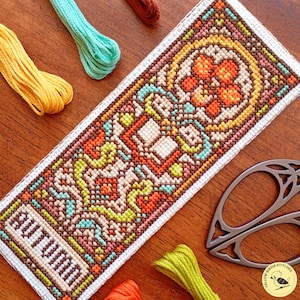 May include: A rectangular cross-stitch bookmark featuring an intricate geometric pattern in shades of orange, brown, yellow, and teal. The word AUTUMN is stitched in brown at the bottom. The piece is surrounded by colorful embroidery floss and scissors.