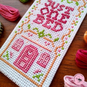 May include: Cross stitch pattern with the text "Just one bed" in pink thread. The pattern is surrounded by a border of orange and green thread.