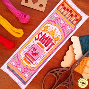 May include: A rectangular cross-stitch bookmark featuring a pink and orange design with the text SHUT UP in bold red letters. The embroidery includes a small jar graphic, decorative floral patterns, and geometric borders on white Aida fabric.
